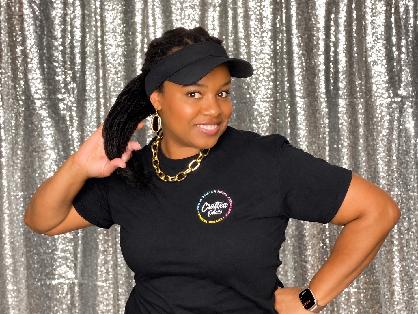 Guest posing in a black CrafTea Details shirt and visor in front of the silver photo booth backdrop, before her AI transformation