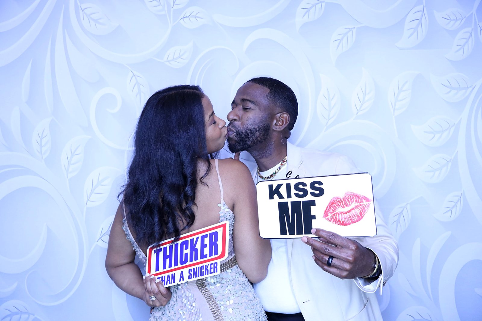 Couple sharing a kiss while holding playful signs in front of a white patterned backdrop