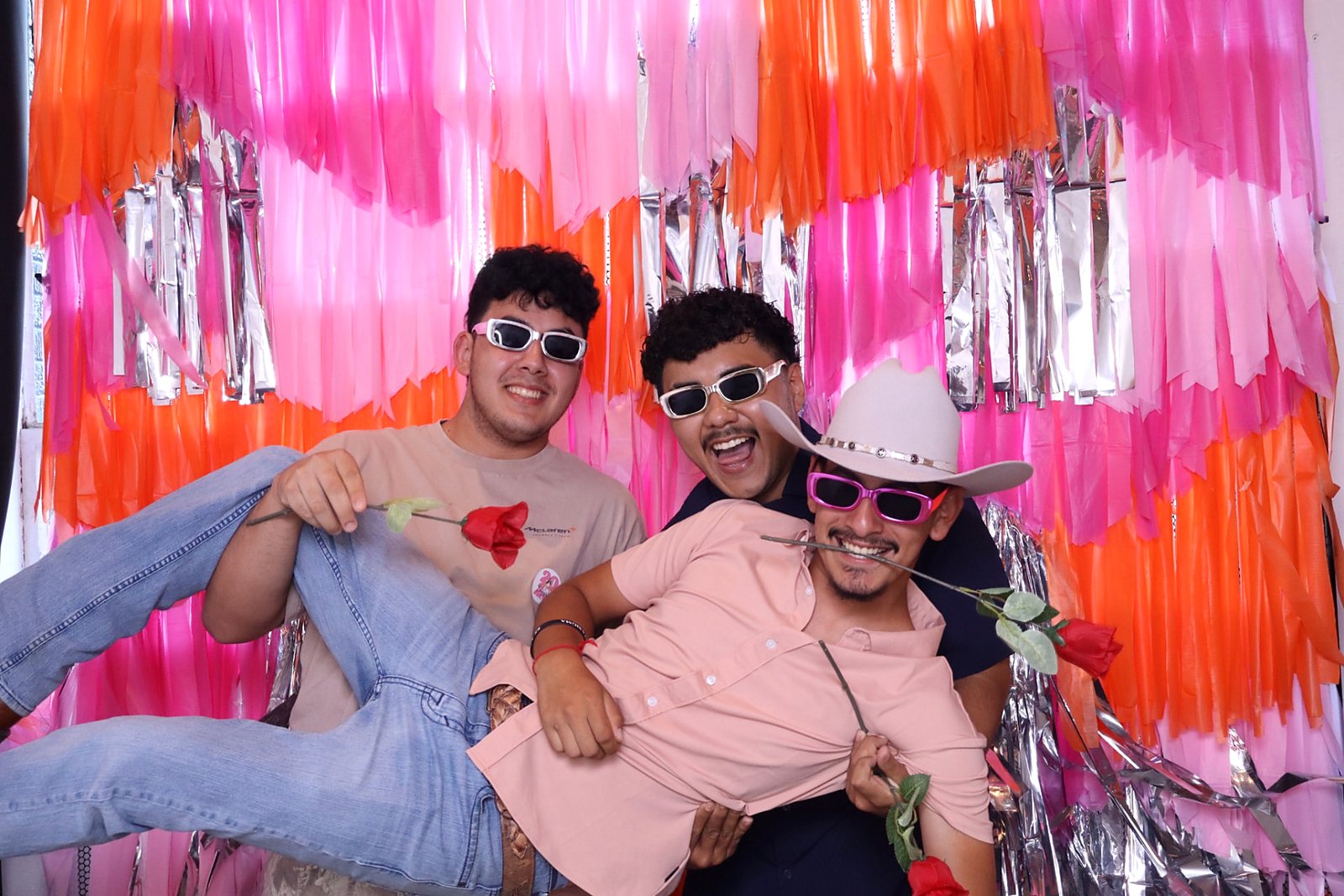 Three guests laughing while holding roses in front of a pink and orange fringe backdrop