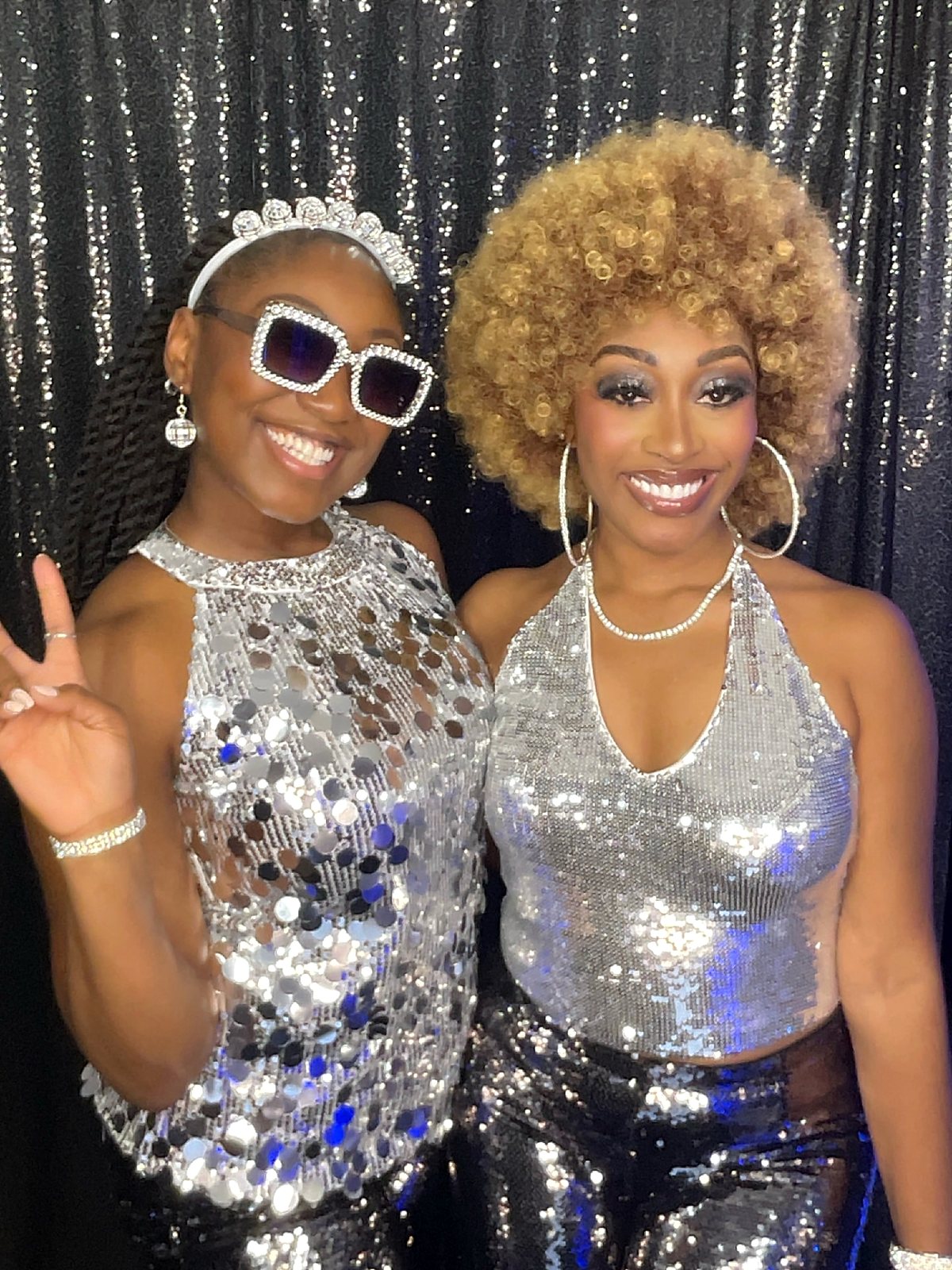 Two guests in silver sequin outfits smiling in front of a black sequin backdrop
